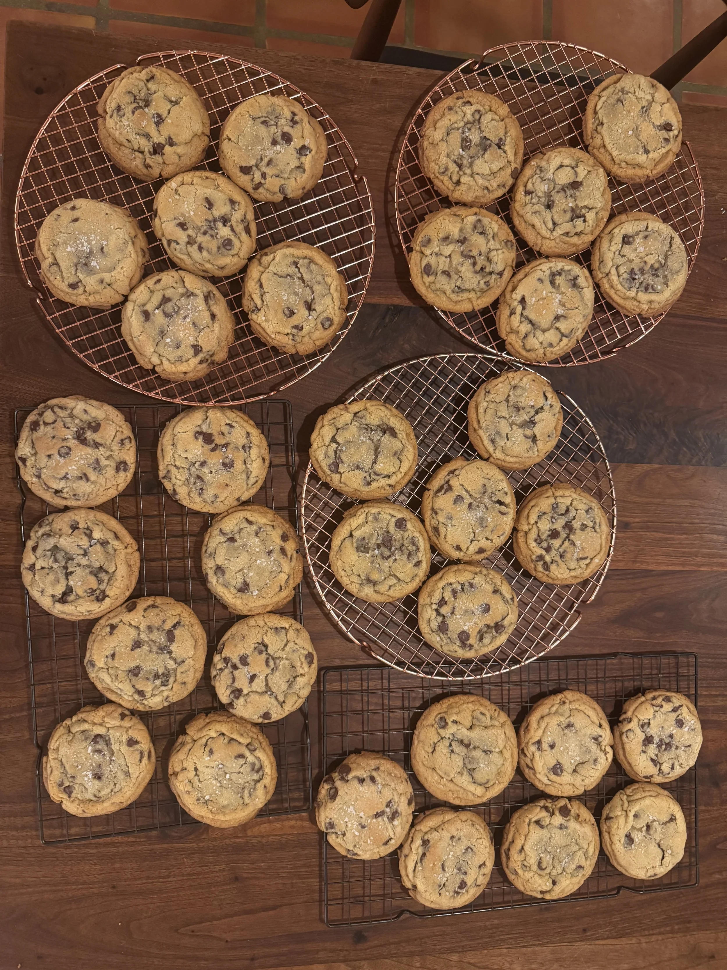 Multiple batches of freshly baked chocolate chip cookies cooling on wire racks placed on a wooden table.