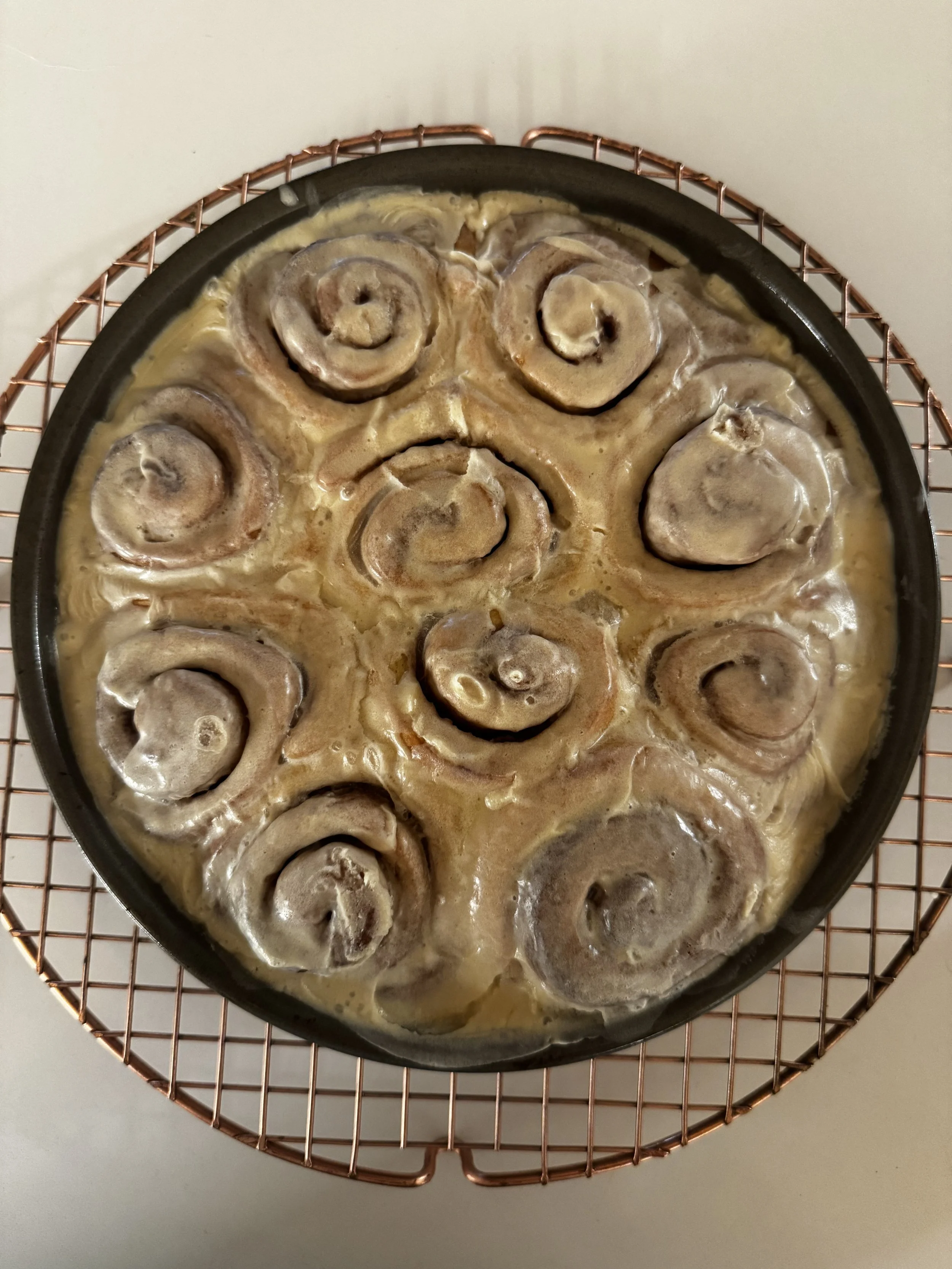 Unbaked cinnamon rolls in a round baking pan filled with frosting, resting on a cooling rack.