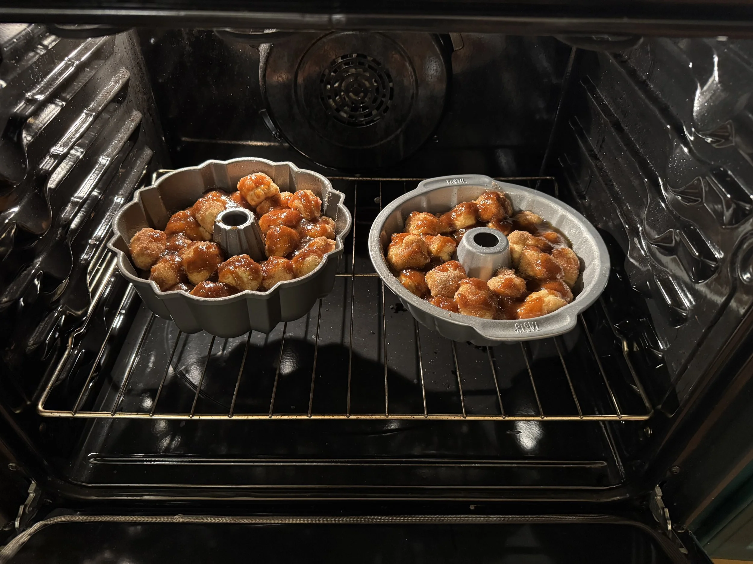 Two Bundt pans filled with small round dough balls coated in glaze inside an oven.