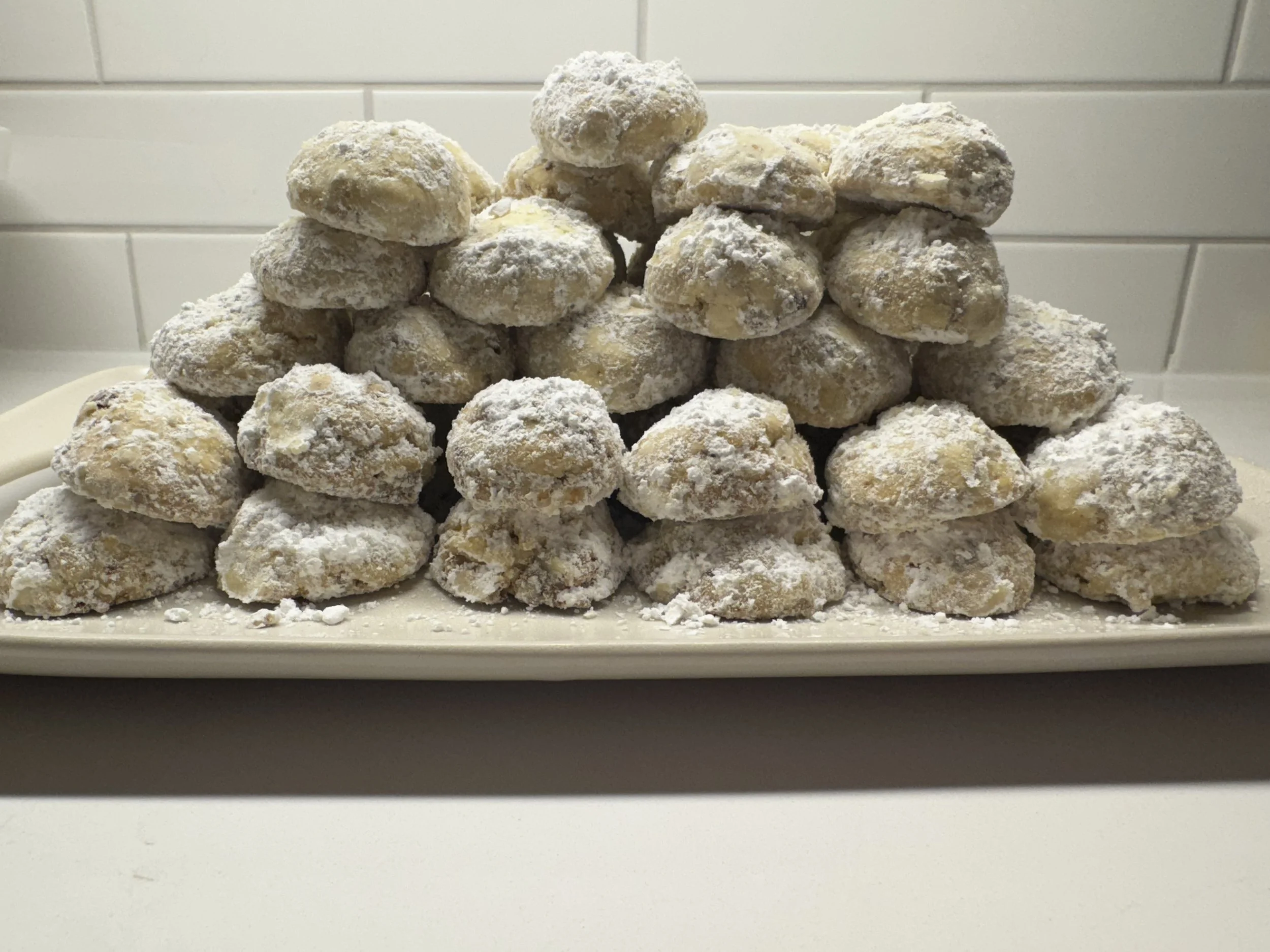 A white rectangular platter holding a pyramid of powdered sugar-coated doughnut holes on a white counter, with a tiled white wall in the background.