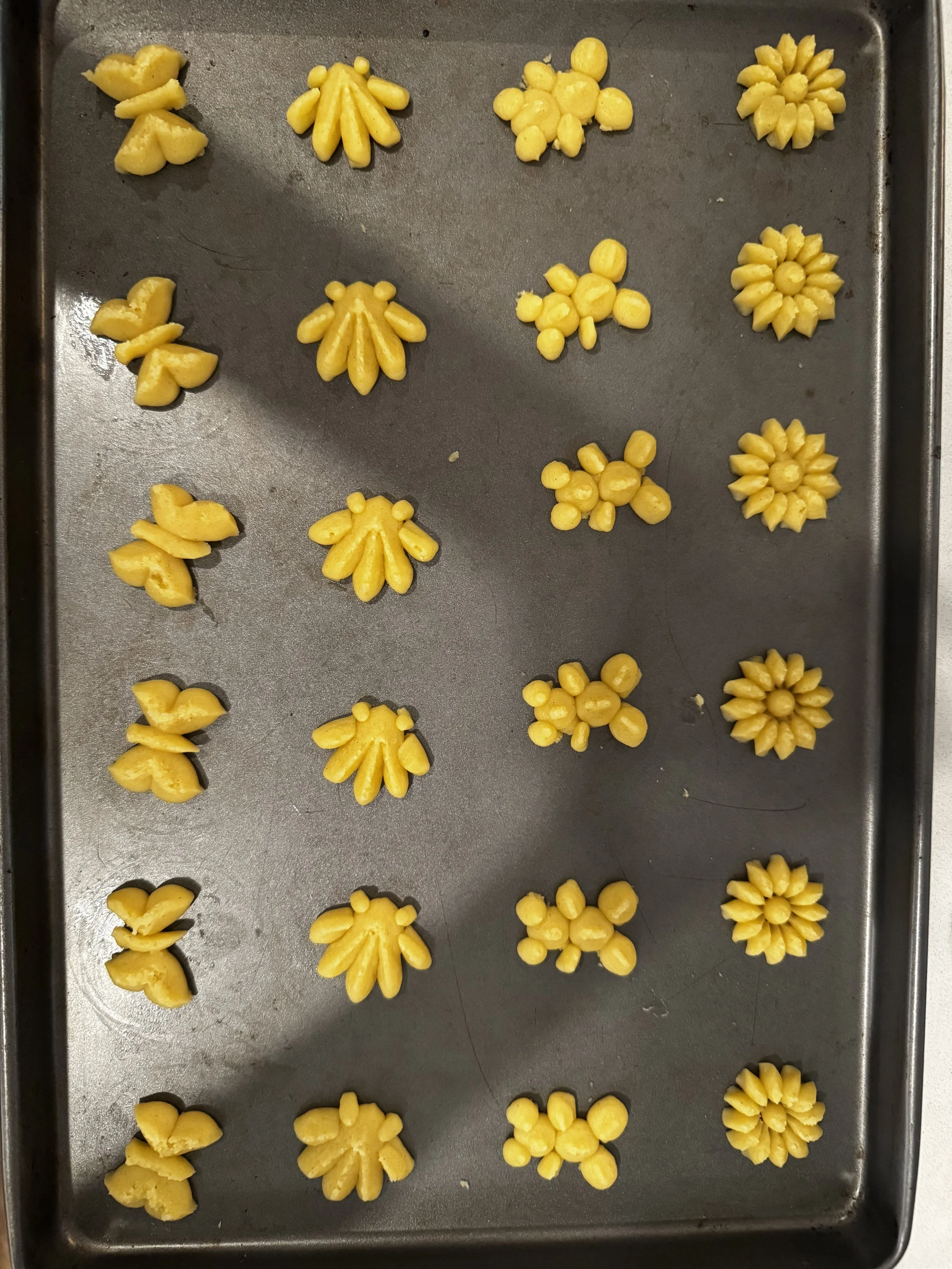 Unbaked decorative yellow cookies shaped like flowers, suns, boats, and other patterns on a metal baking tray.