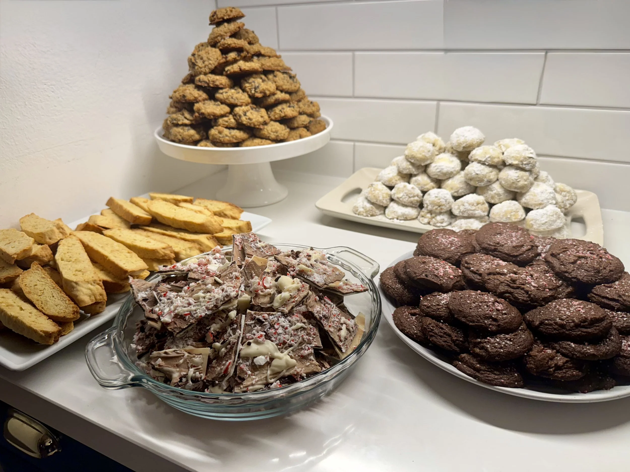 An assortment of Christmas cookies and treats on a white table, including chocolate cookies, powdered sugar snowball cookies, peppermint bark, biscotti, and a stack of small chocolate chip cookies.