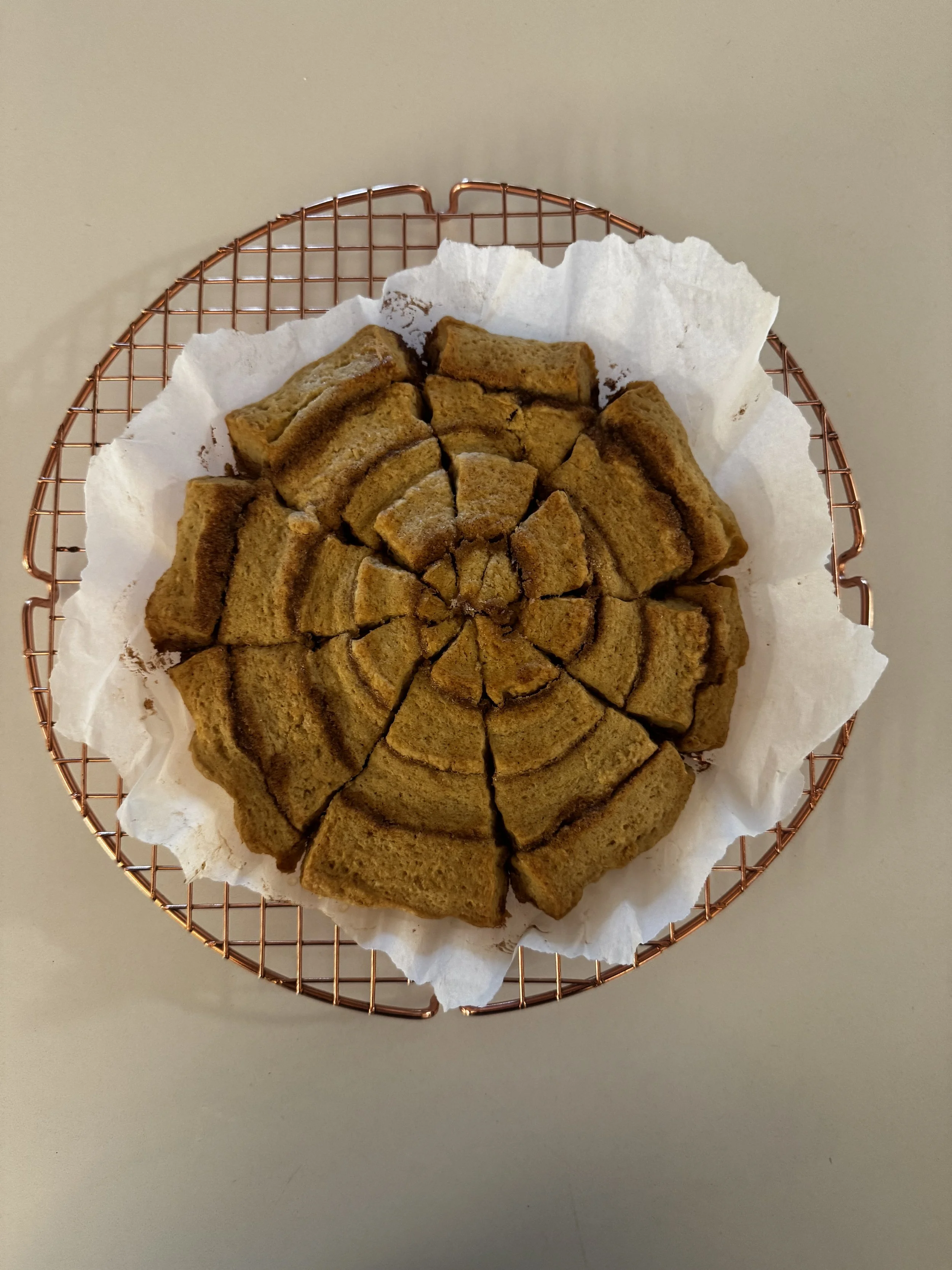 A round stack of sliced, homemade cinnamon bread with a golden-brown crust, placed on white parchment paper inside a copper wire cooling rack.