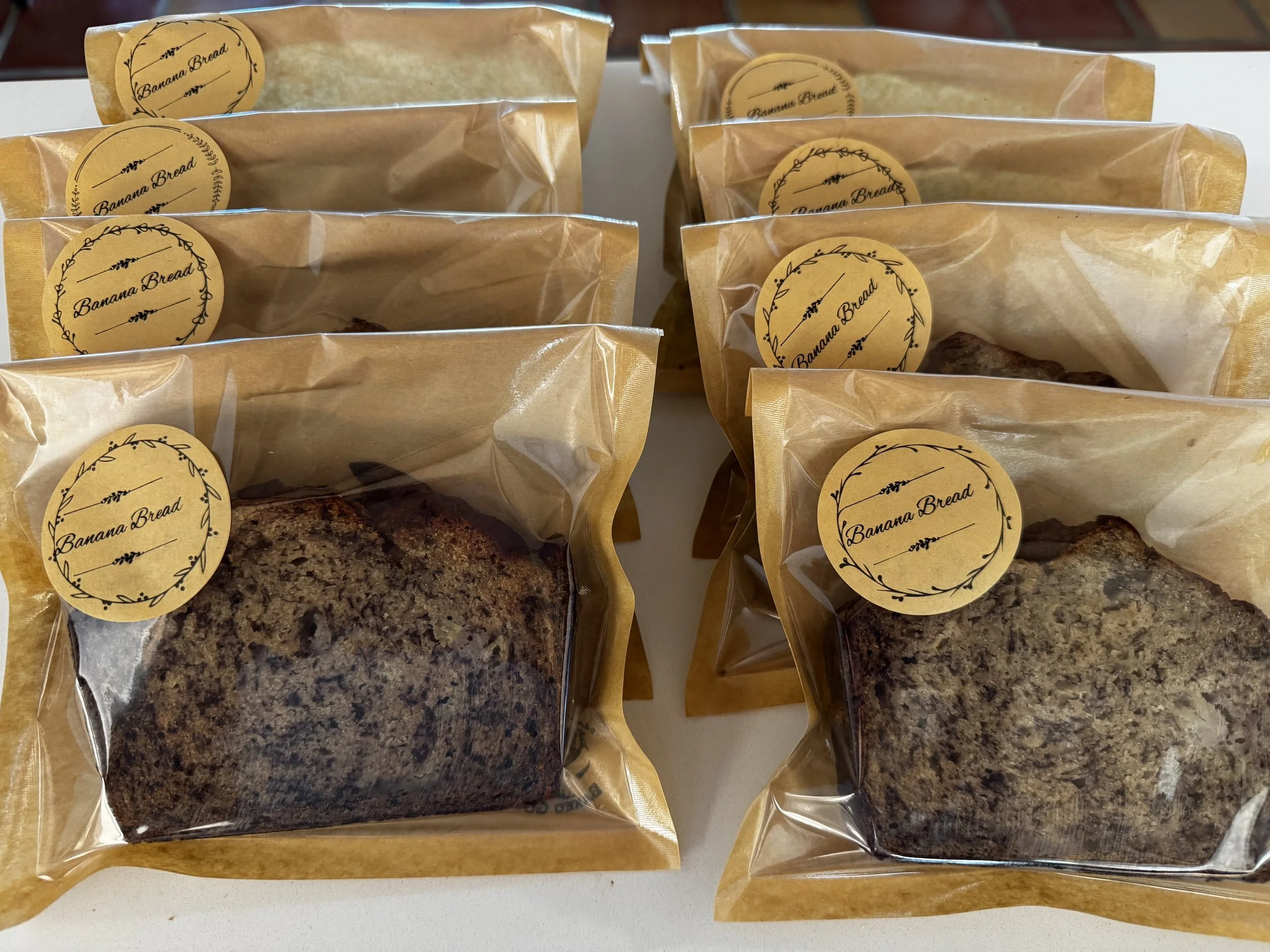 Several bread loaves in clear plastic packaging with a yellow label reading "Banana Bread" arranged on a white surface.