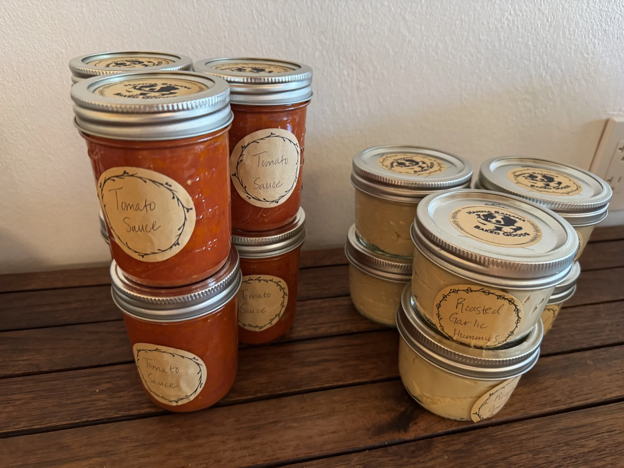 Several glass jars of homemade tomato sauce and roasted garlic hummus with handwritten labels on a wooden surface.