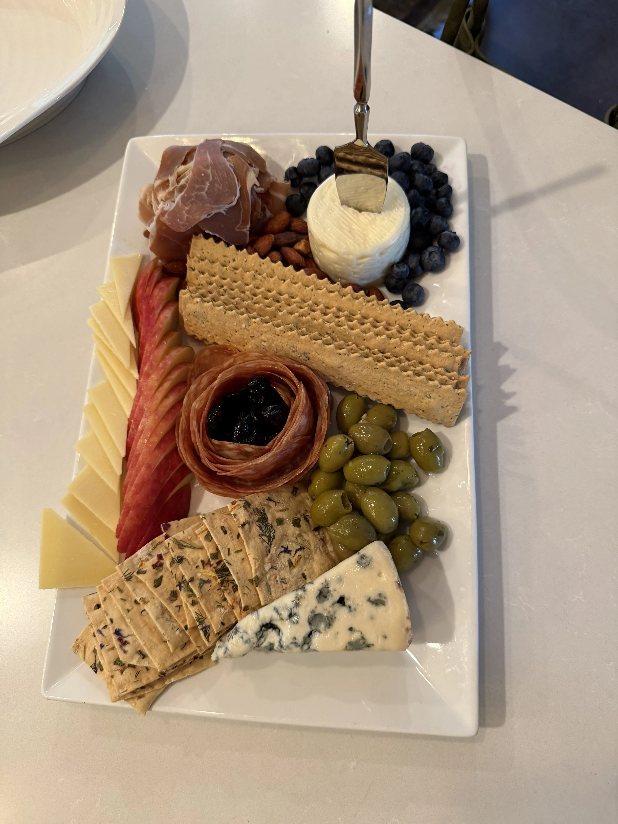 Cheese platter with various cheeses, sliced apples, green olives, prosciutto, almonds, crackers, blue cheese, and blueberries.