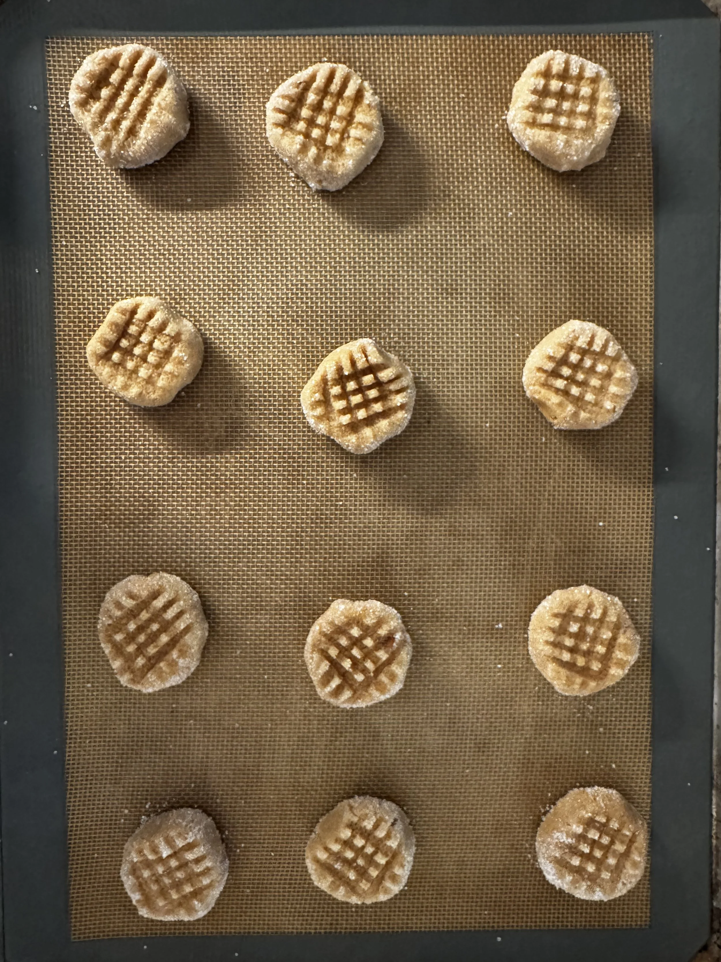A baking sheet with 12 unbaked cookies, arranged in three rows of four, on a silicone baking mat. The cookies are round and have a crisscross pattern on top.