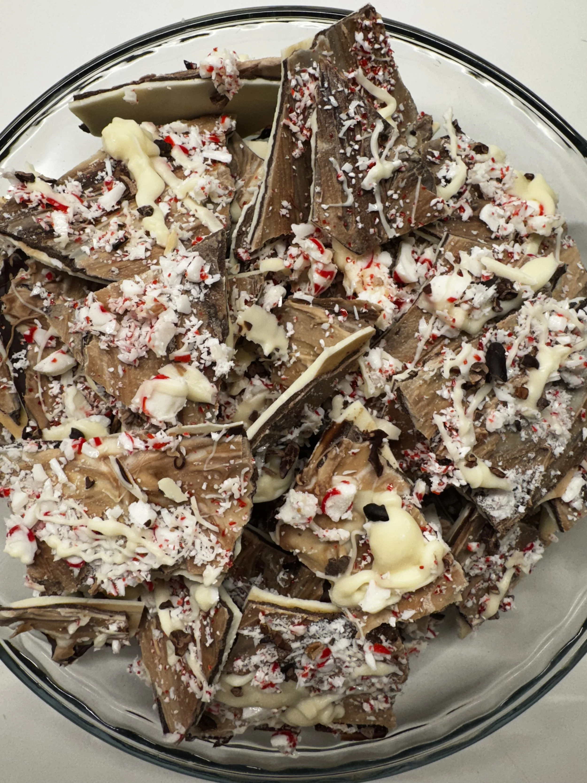 A glass bowl filled with broken chocolate bark topped with crushed peppermint, white chocolate drizzle, and coconut flakes.
