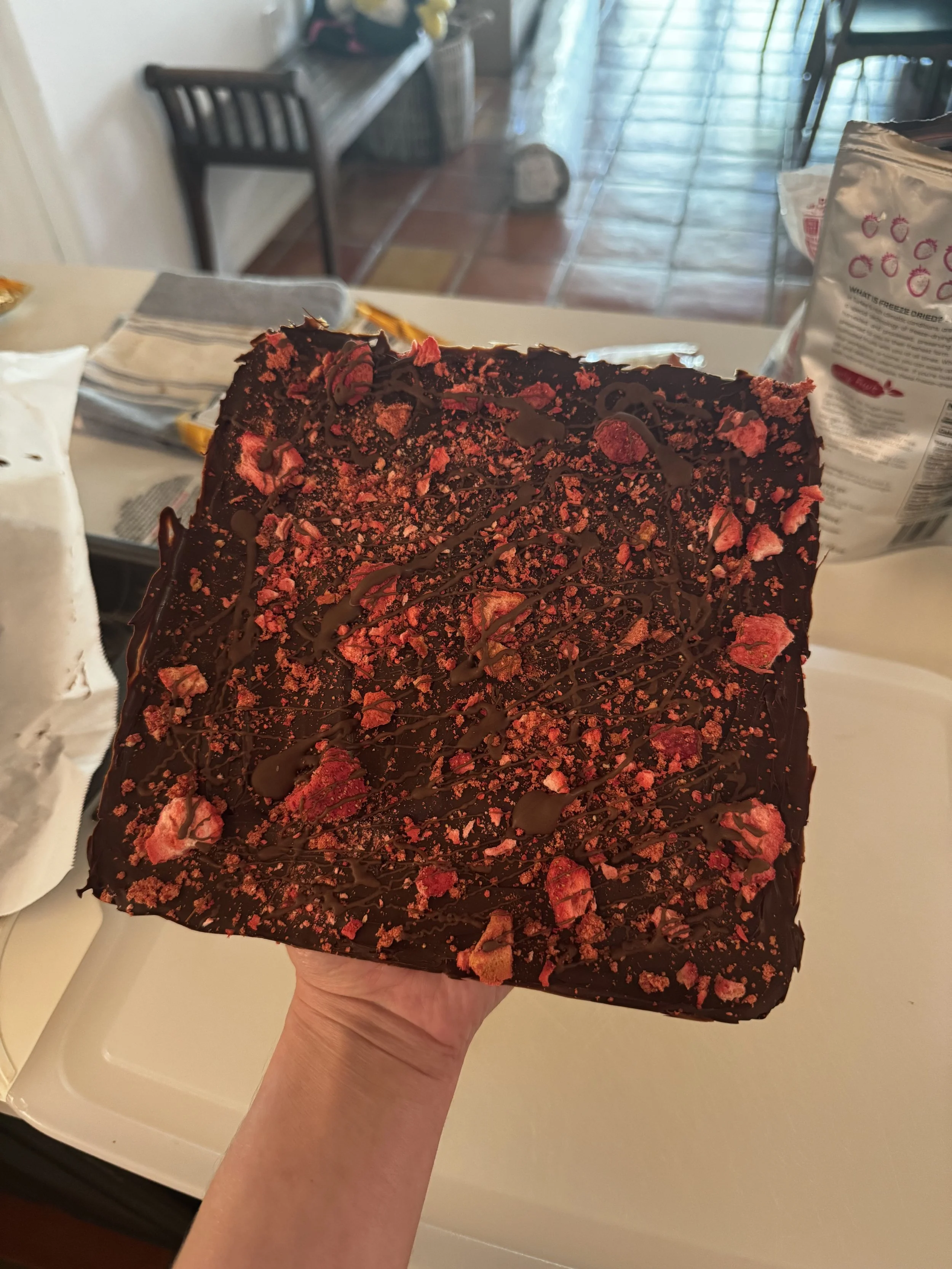 A person holding a large square chocolate dessert topped with crushed strawberries and drizzled with chocolate sauce.