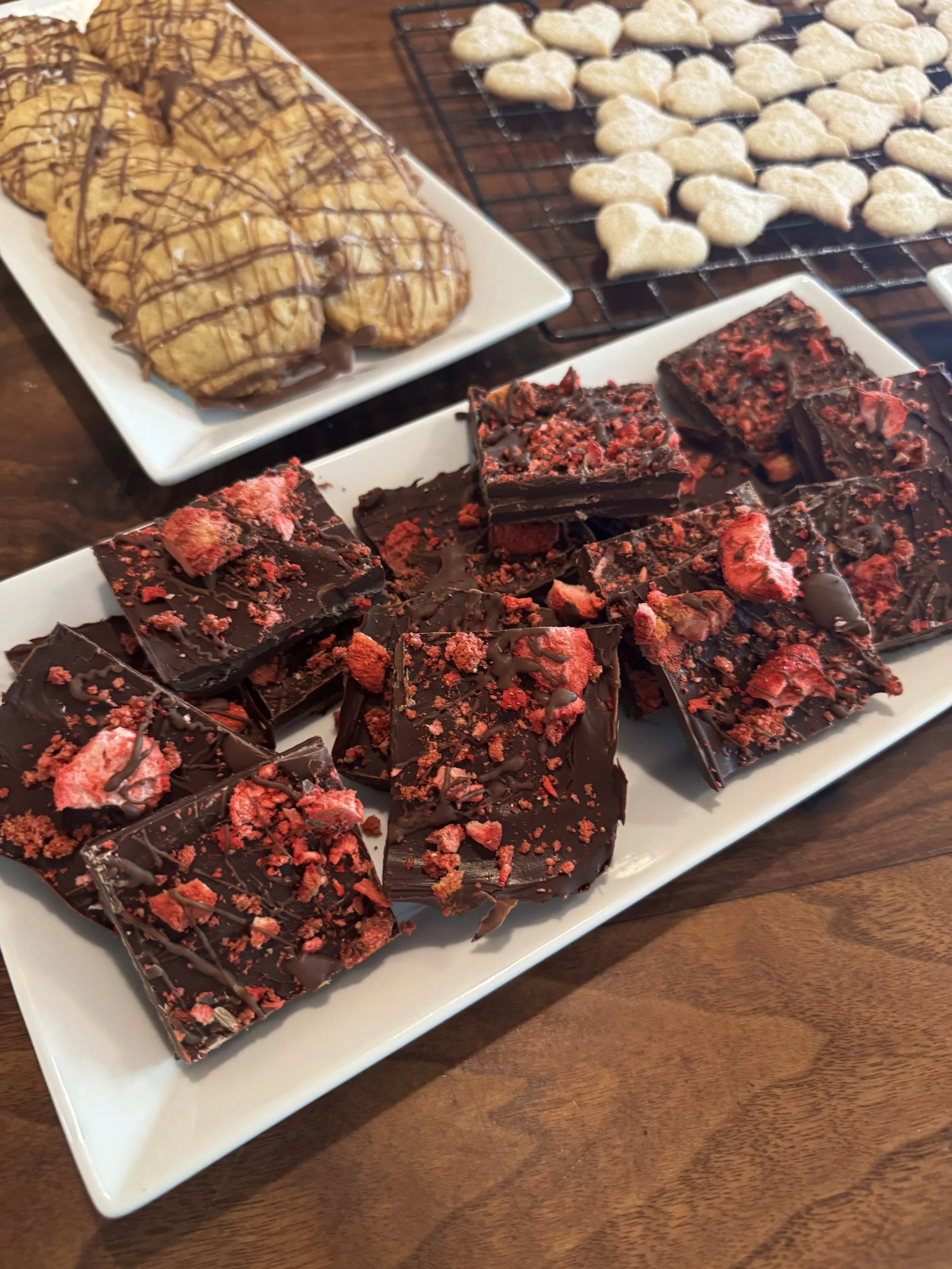 Various homemade cookies and chocolate bark on white plates and a cooling rack on a wooden table.