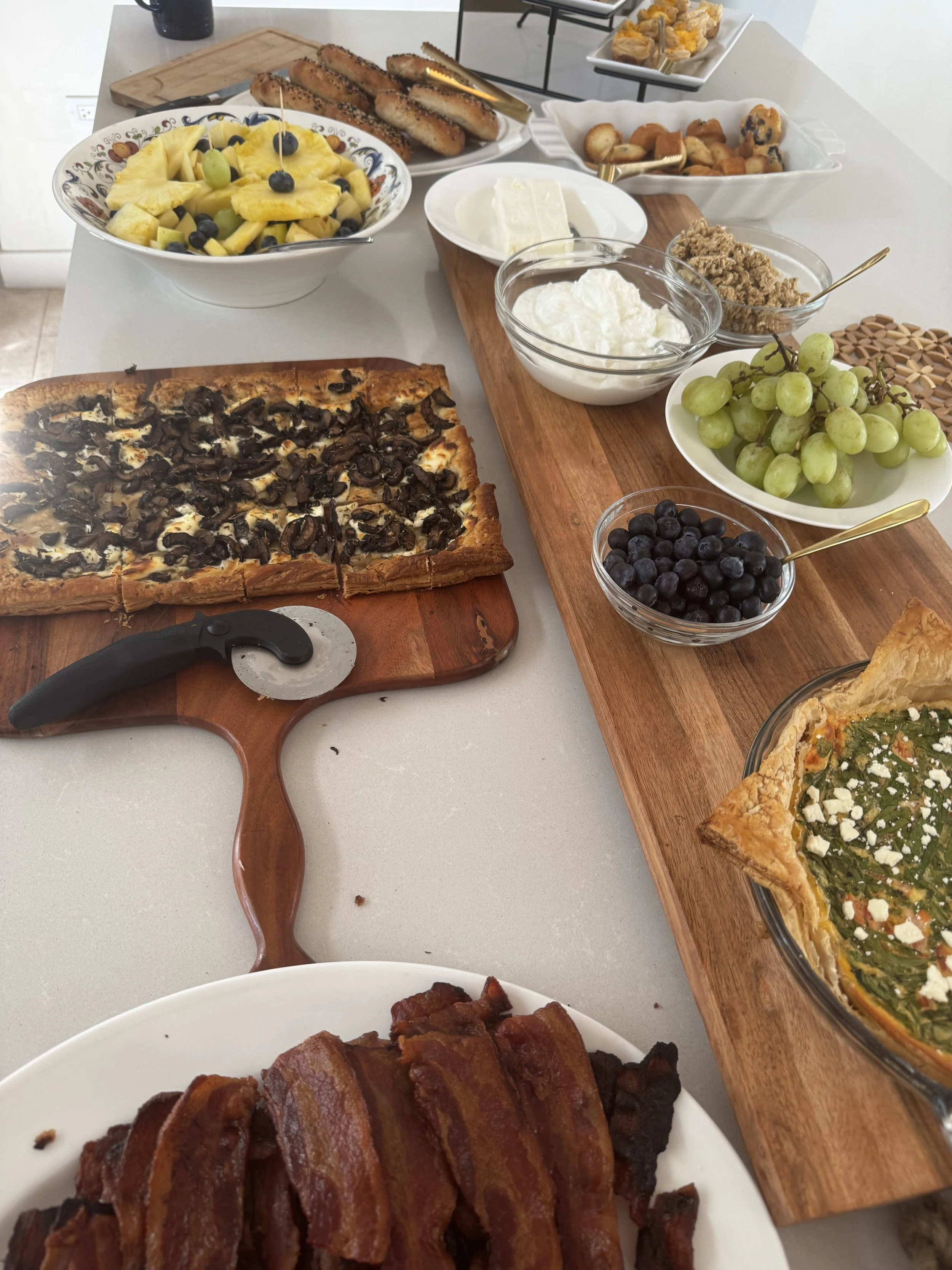 Table with breakfast foods including bacon, pineapple and blueberry fruit salad, mushroom quiche, veggie quiche, fresh grapes, blueberries, whipped cream, cream cheese, breadsticks, and pastries.