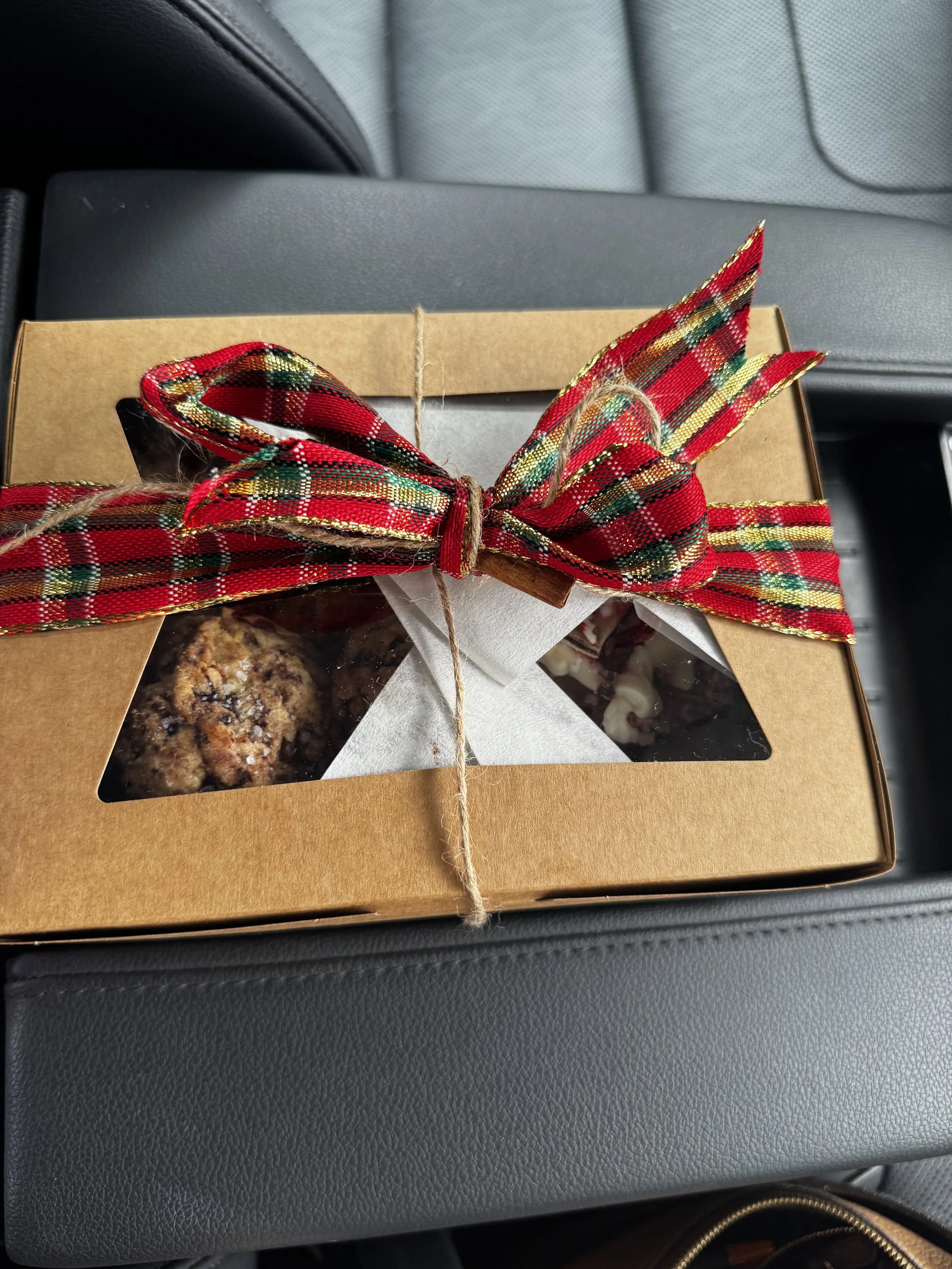 A cardboard gift box with a clear plastic window, decorated with a red, green, and gold tartan ribbon bow, resting on a car seat. Inside the box, cookies and chocolates are visible.