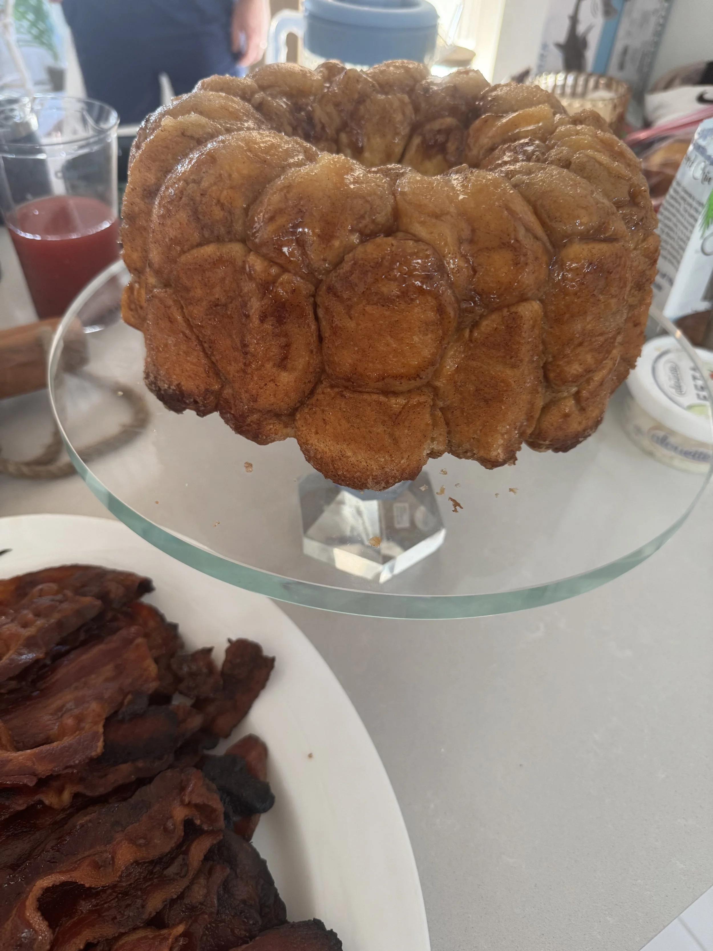 A large, battered and fried apple fritter on a glass cake stand, with bacon and drinks in the background.