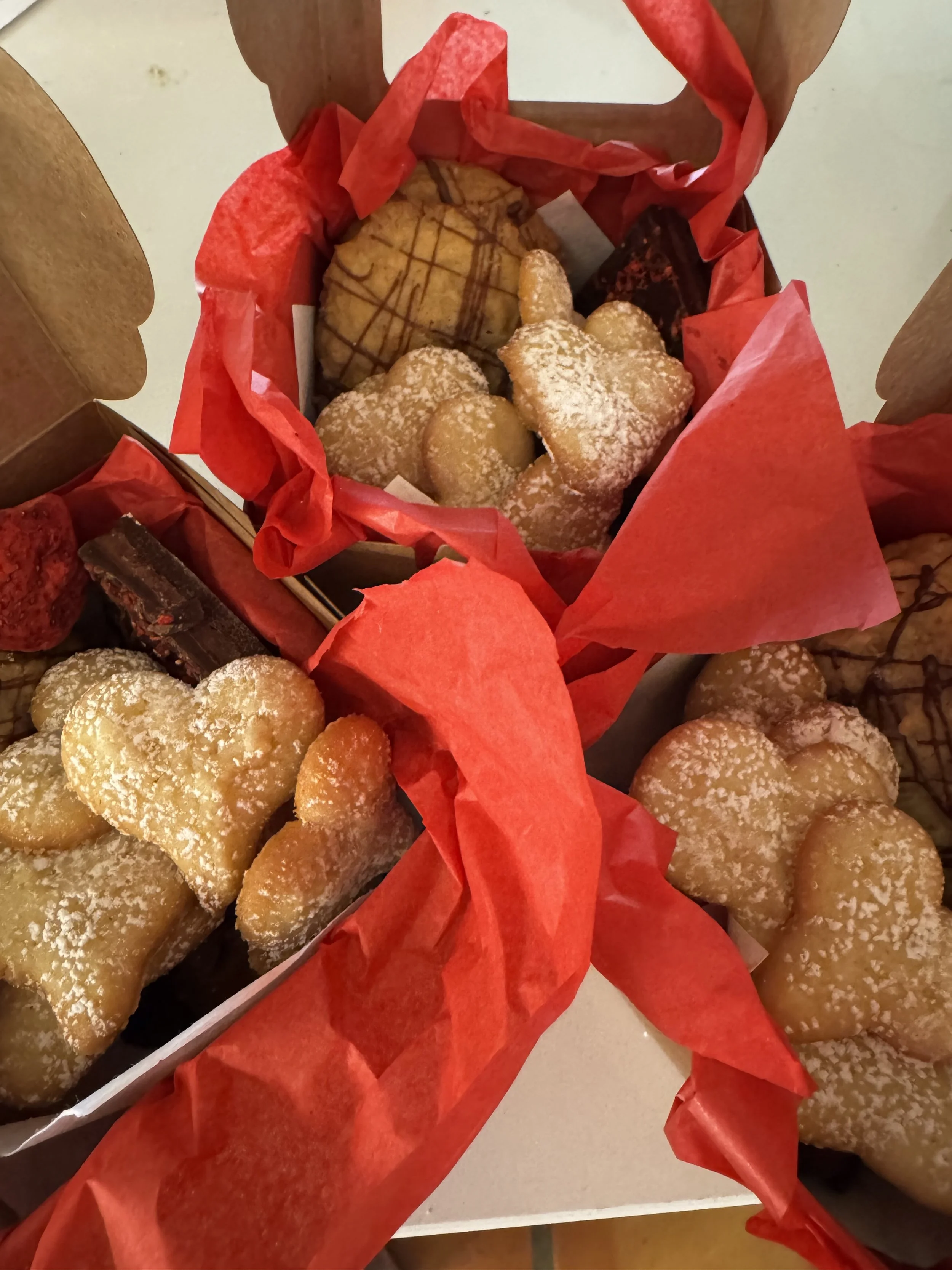 Assorted cookies, including heart-shaped and rectangular cookies dusted with powdered sugar, in red gift boxes lined with red tissue paper.