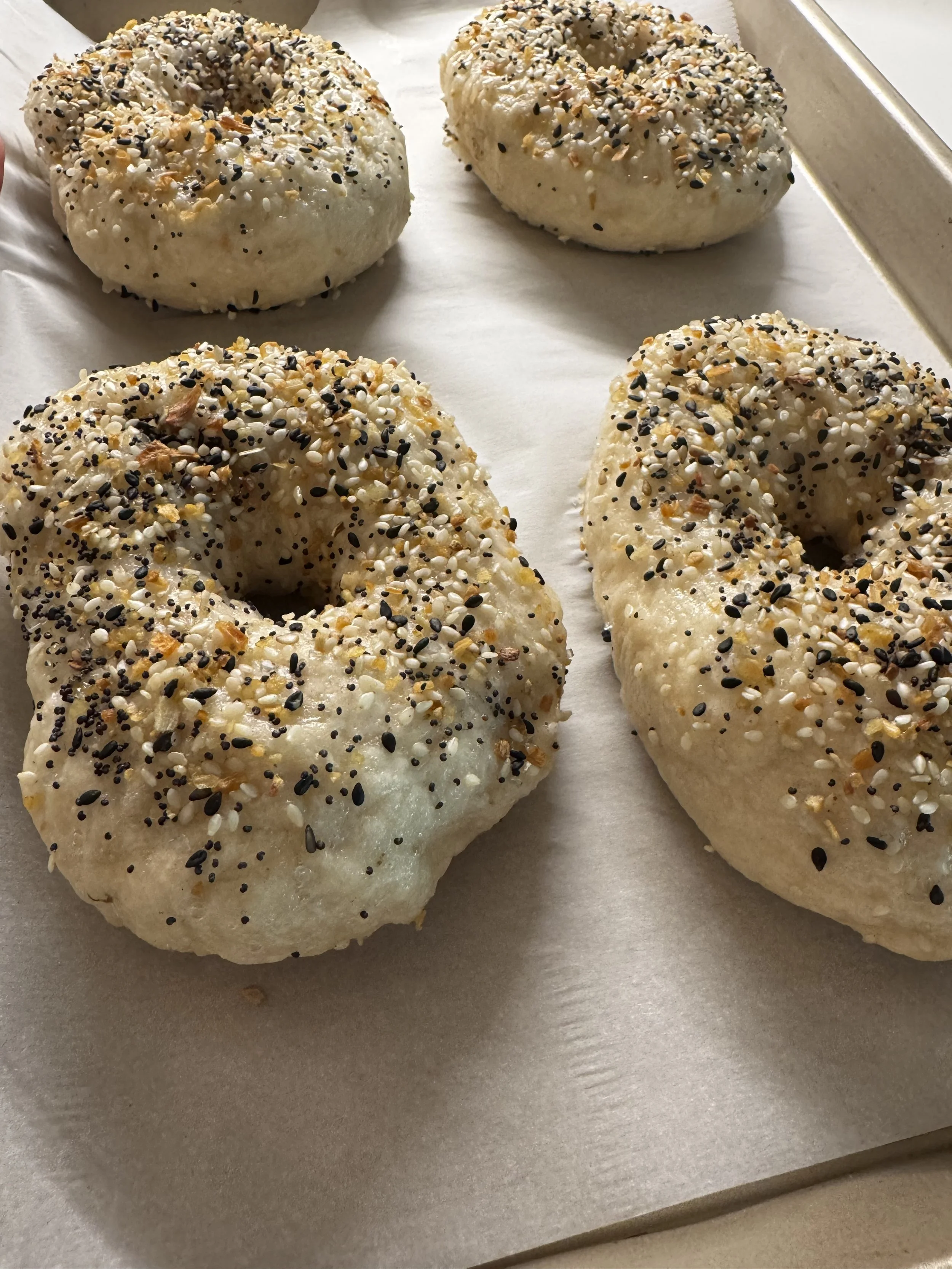 Four bagels on a baking sheet, topped with mixed seeds and spices, ready for baking or cooling.