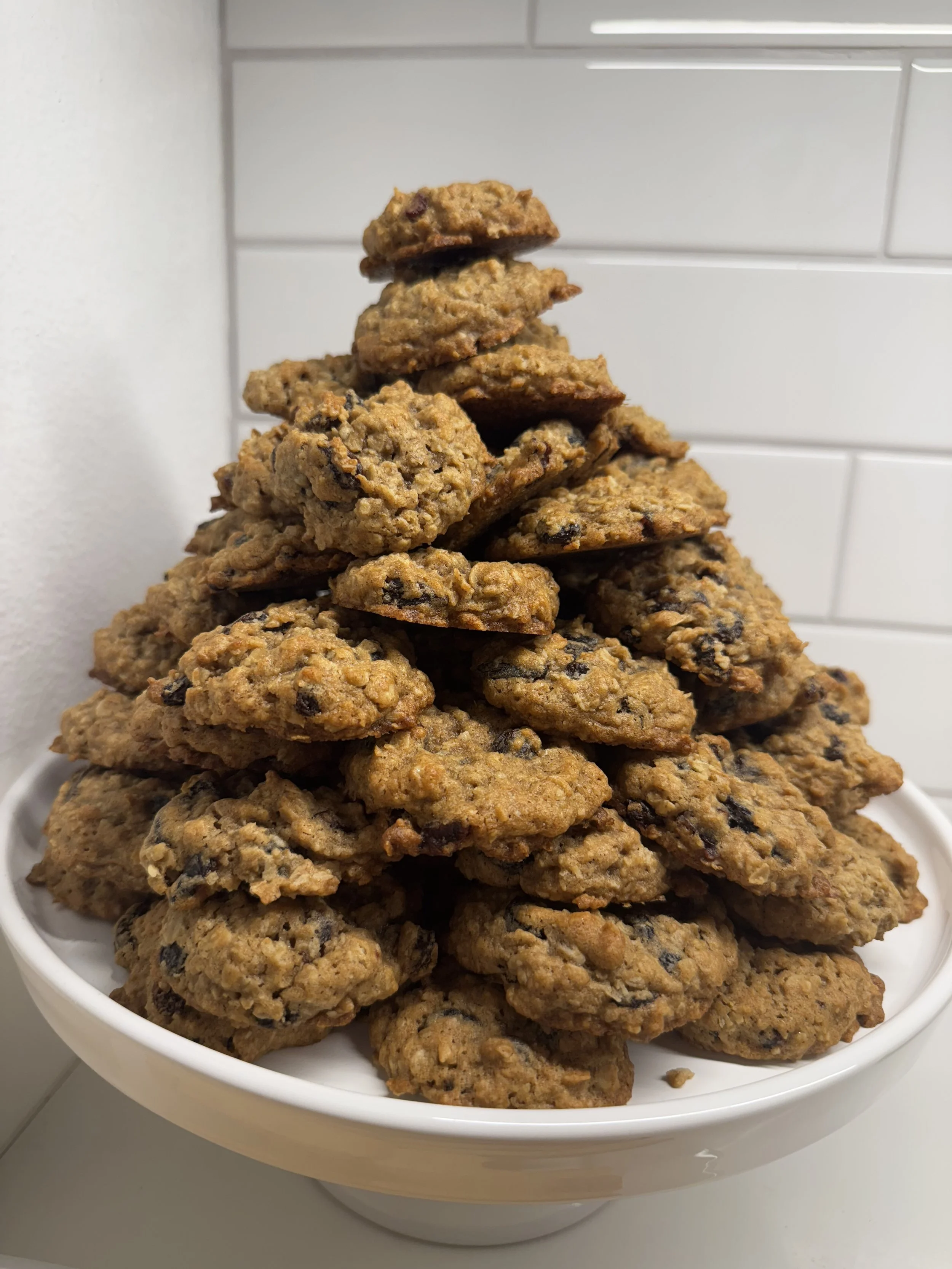 A pyramid-shaped stack of chocolate chip cookies on a white plate against a white tiled background.