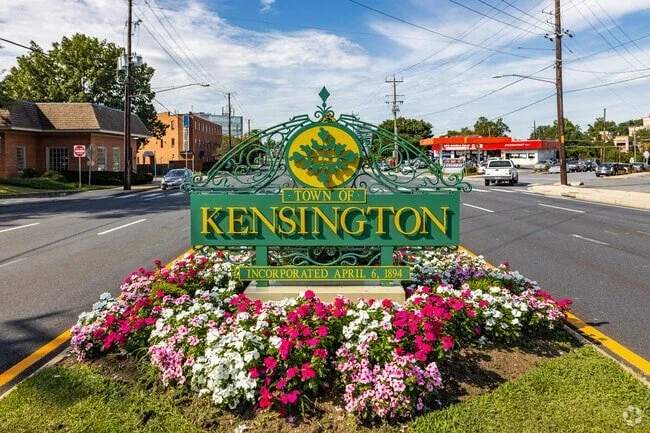 A decorative sign for the town of Kensington surrounded by a colorful flower bed with pink, white, and purple flowers, located at an intersection on a sunny day.
