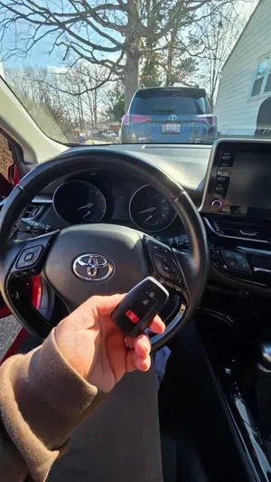 Inside a car, person holding car key fob, dashboard, steering wheel, and an outside view of parked vehicles and trees.