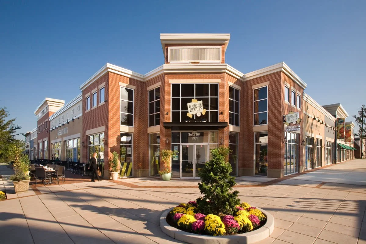 A shopping mall with a brick exterior, large glass windows, and an entrance marked 'Suite 104 5331'. There are outdoor tables and chairs, plants, and a flower bed with yellow and pink flowers in the foreground.
