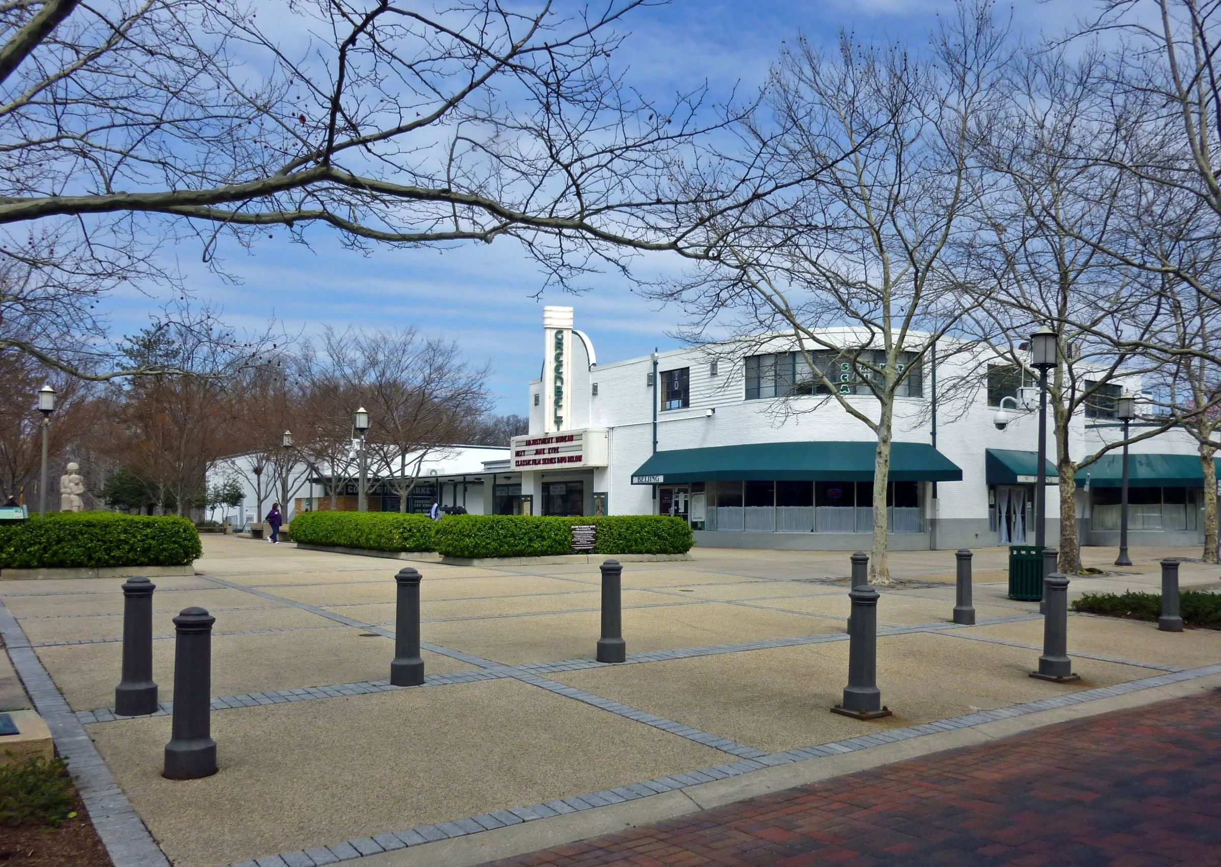 A small outdoor shopping area with a large white building, old-fashioned street lamps, leafless trees, and a paved plaza with black bollards.
