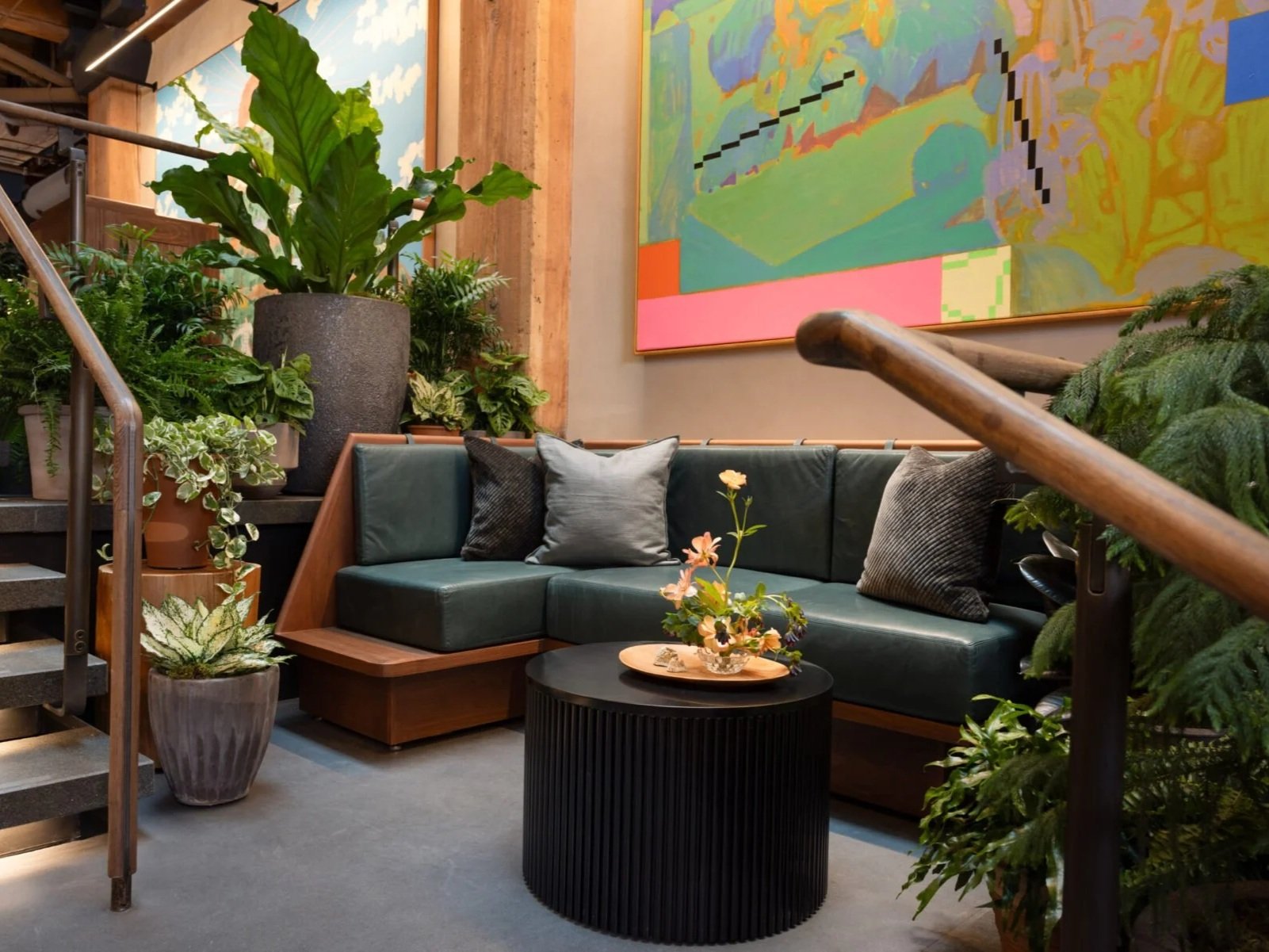 Indoor seating area with a dark green sectional sofa, multiple pillows, a black round coffee table with a floral arrangement, and numerous potted plants, with colorful artwork on the wall behind.