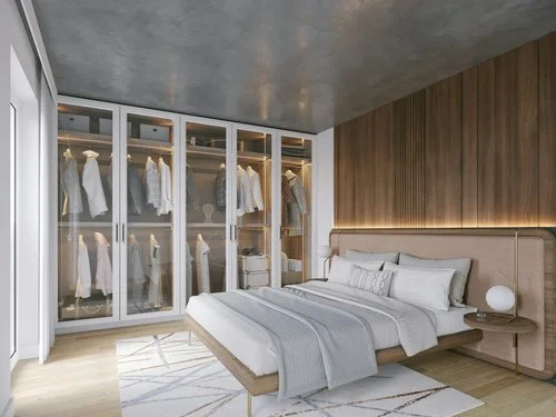 Modern bedroom with a large bed, white bedding, and an upholstered headboard. Beside the bed is a round white bedside lamp and a wooden accent wall. There is a glass closet with white robes and shelves.