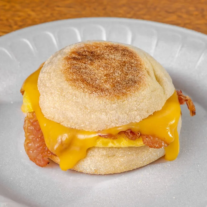 Close-up of a breakfast sandwich with biscuits, cheddar cheese, scrambled eggs, and bacon on a white disposable plate.