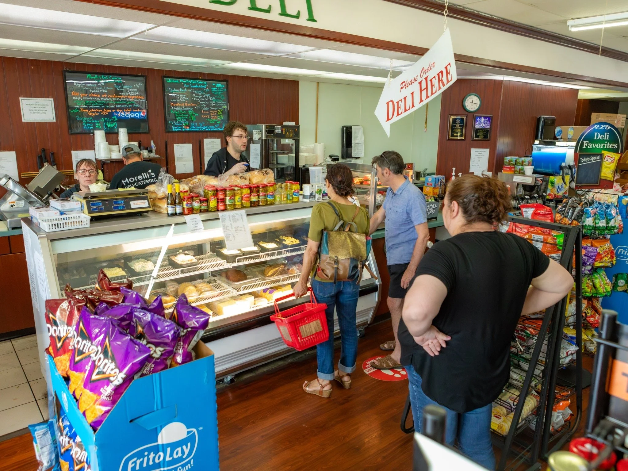 Inside a deli with several customers waiting in line to order food. There is a display case with baked goods and snacks, with a person behind the counter preparing orders. A sign hanging above says 'Please order Deli Here'.