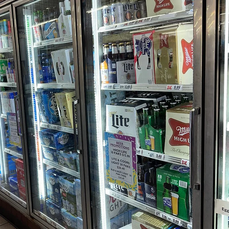 A supermarket fridge filled with various brands of beer, including Miller, Bud Light, and Busch, with prices and promotional signs.