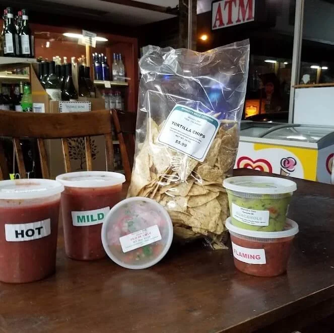 Small table with plastic cups of salsa labeled hot, mild, and flaming, a container of guacamole, a container of salsa, a bag of tortilla chips, and a bowl of pico de gallo. Background shows shelves with liquor bottles and an ATM sign.