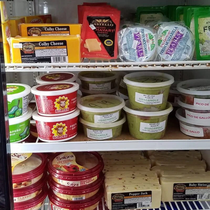 Refrigerator shelves stocked with cheese and dip containers, including guacamole, red pepper, and Swiss cheese.