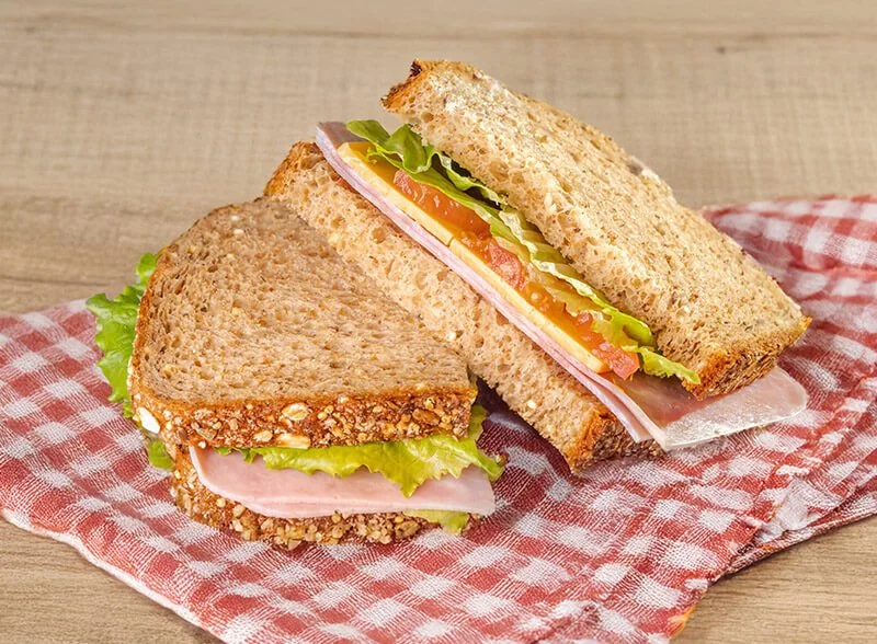 Close-up of two whole grain and one sliced sandwich with lettuce, tomato, turkey, and cheese on a red and white checkered cloth.