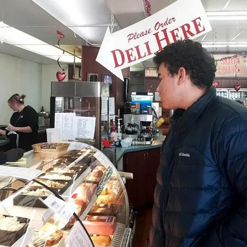 A man in a black jacket is looking at a deli display case filled with various meats and cheeses inside a deli or cafe. A sign above reads 'Please Order Deli Here.' A woman with pink hair is working behind the counter in the background.
