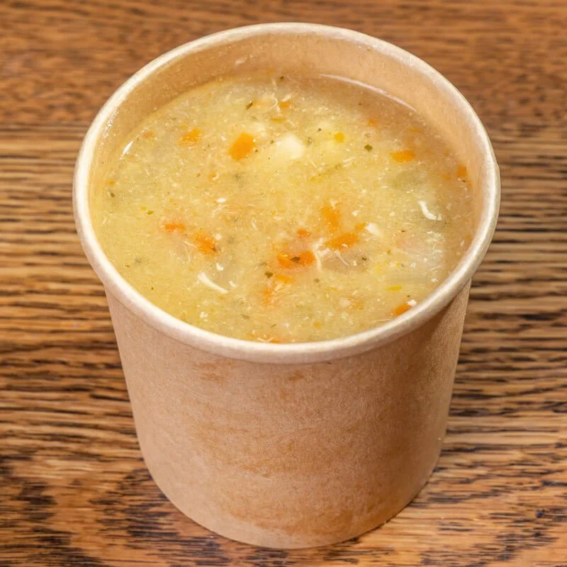 A paper cup filled with creamy soup containing small pieces of vegetables, placed on a wooden surface.