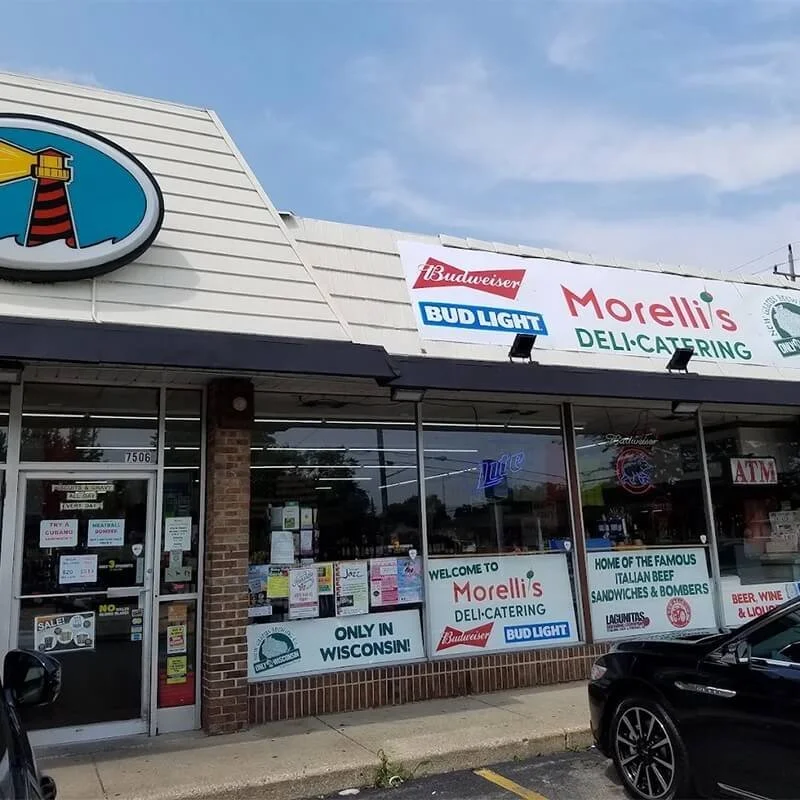 Exterior view of a deli-catering shop named Morelli's, with signs promoting Budweiser and Bud Light, and windows displaying various notices and food advertisement posters.