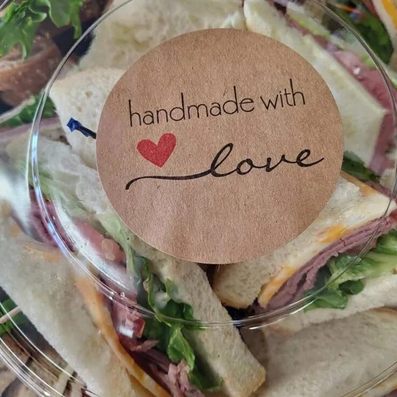 A clear plastic container holding sandwich triangles with layers of ham, cheese, lettuce, and bread, topped with a round paper label that says 'handmade with love' with a red heart.