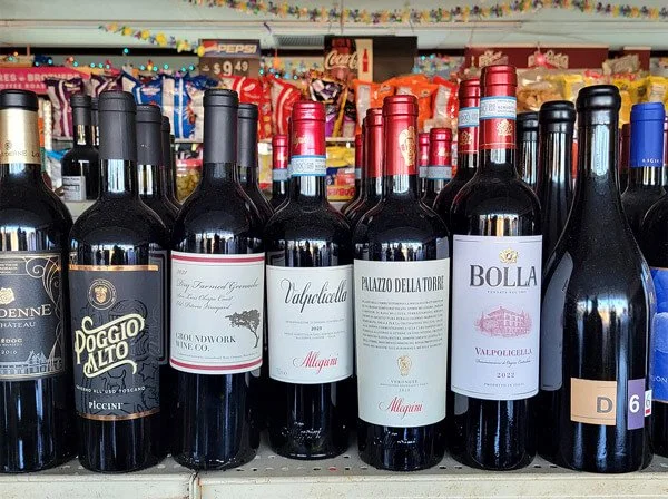 A row of seven bottles of red wine on a store shelf, with various labels and caps, behind snacks and candy.