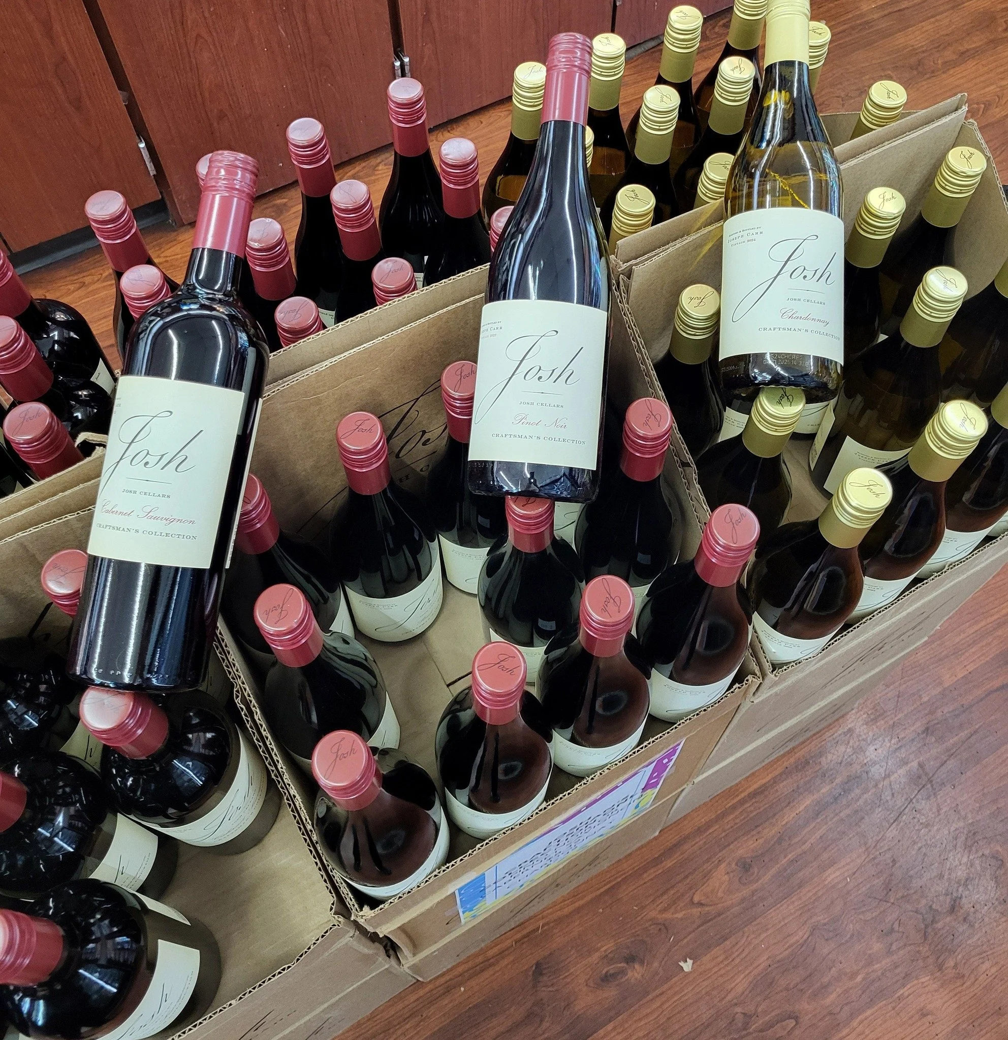 Box of bottles of Josh wine, including Cabernet Sauvignon, Pinot Noir, and Chardonnay, with red and yellow caps, on a wooden floor.