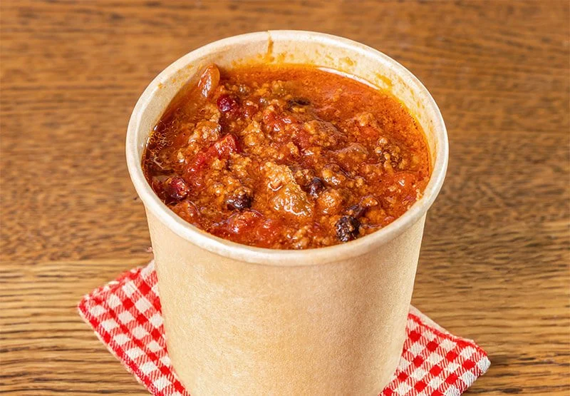 A paper cup of chili with ground beef, beans, and tomato sauce on a wooden surface with a red and white checkered napkin.