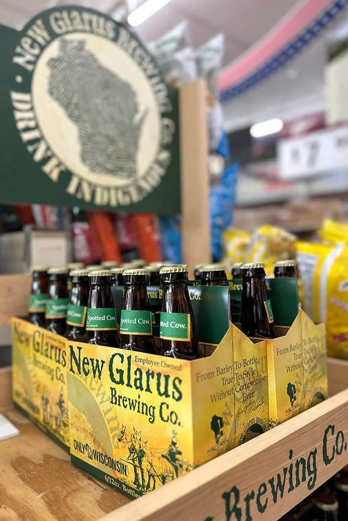 A cardboard carrier holding bottles of beer from New Glarus Brewing Co., with a sign in the background featuring the company's logo.