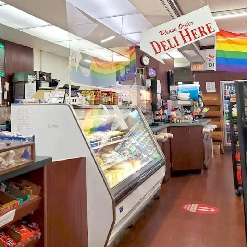 Interior of a grocery store with a refrigerated display case, checkout counter, and a rainbow flag decoration on the wall. A sign hanging from the ceiling says "Please Order Deli Here."