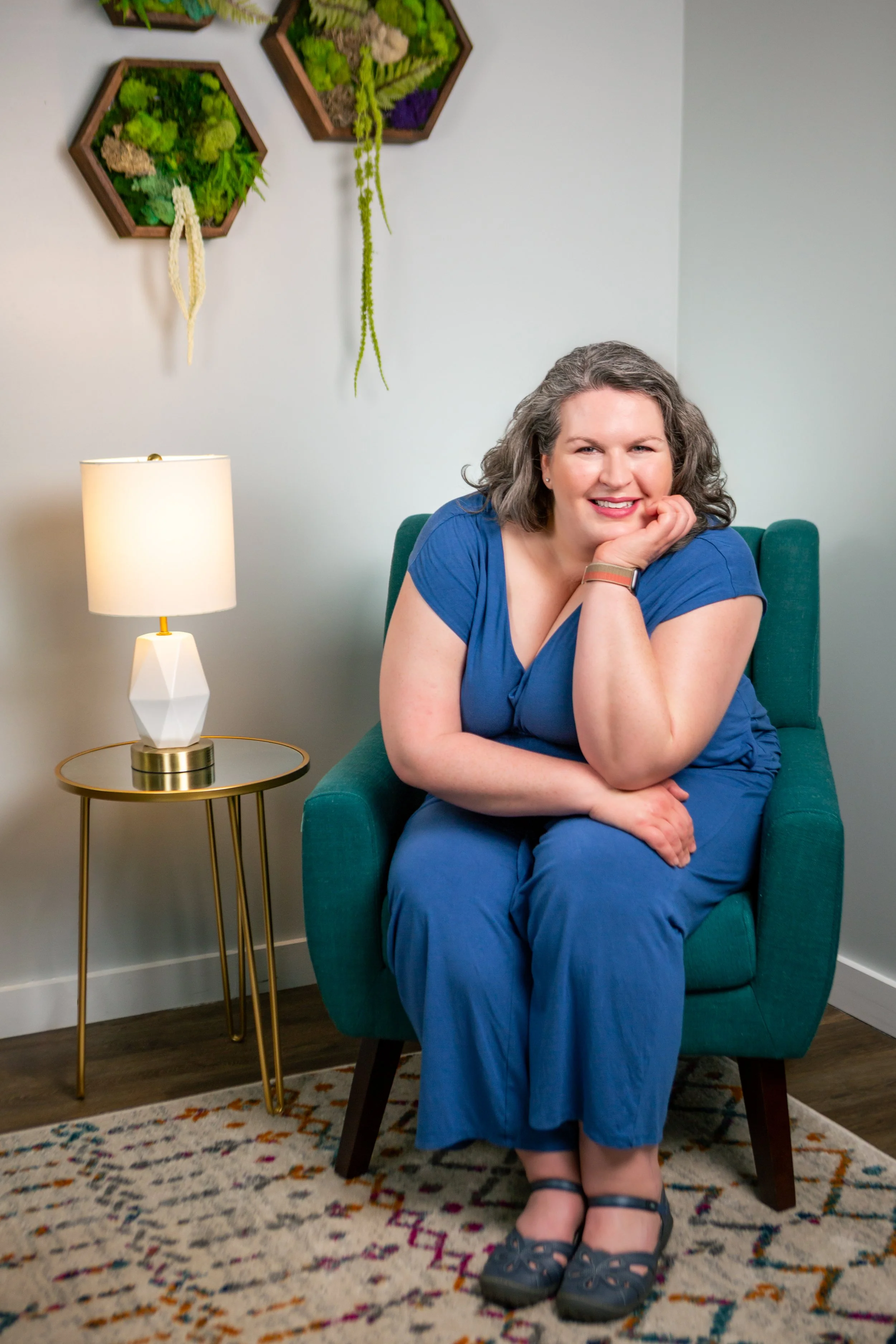 A woman with curly gray hair, wearing a blue dress and black sandals, sitting on a teal armchair in a living room, smiling with her head resting on her left hand. There is a white lamp with a gold base on a side table next to her, and wall art with hexagonal planters filled with plants hanging on the white wall behind her. A patterned rug is visible under her feet.