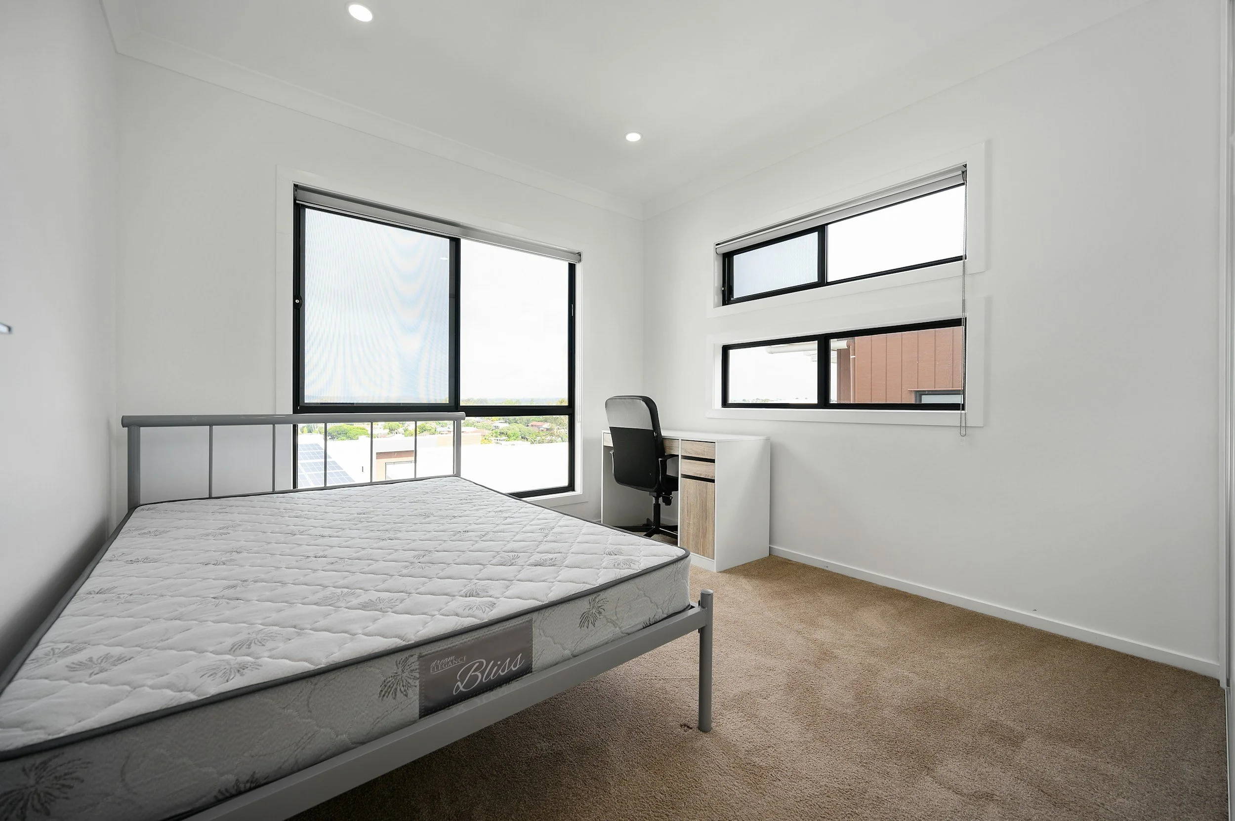 Empty bedroom with a metal bed frame and a mattress, a small white desk with a black office chair, large windows with blinds, beige carpet, white walls, and recessed ceiling lights.