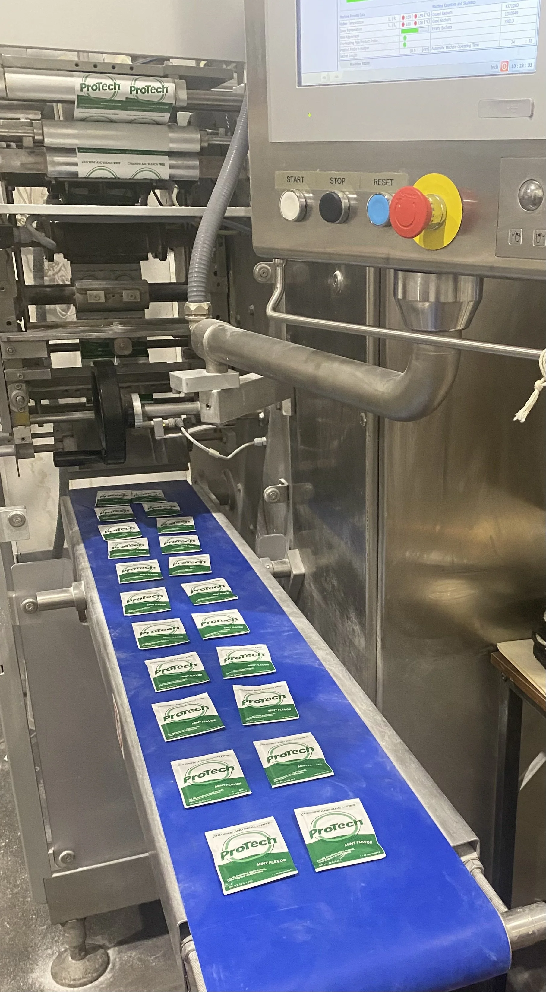 A manufacturing line with a conveyor belt carrying small packets of Protech mint-flavored cleaner for oral removables, and a machinery control panel with buttons and a screen.