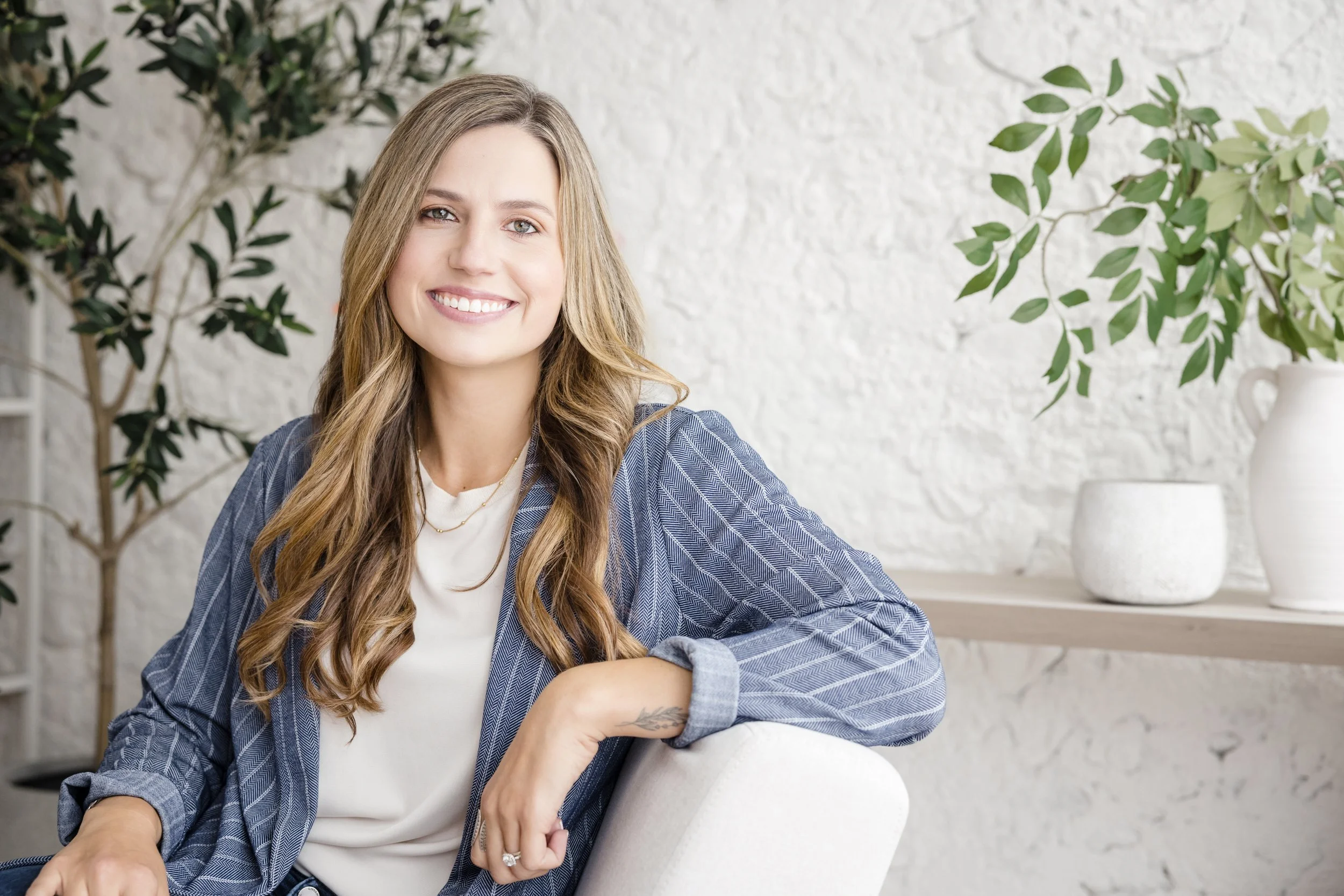 A smiling woman with long light brown hair sitting on a white chair in a bright space with a white textured wall, green leafy plants, and white decorative objects in the background.