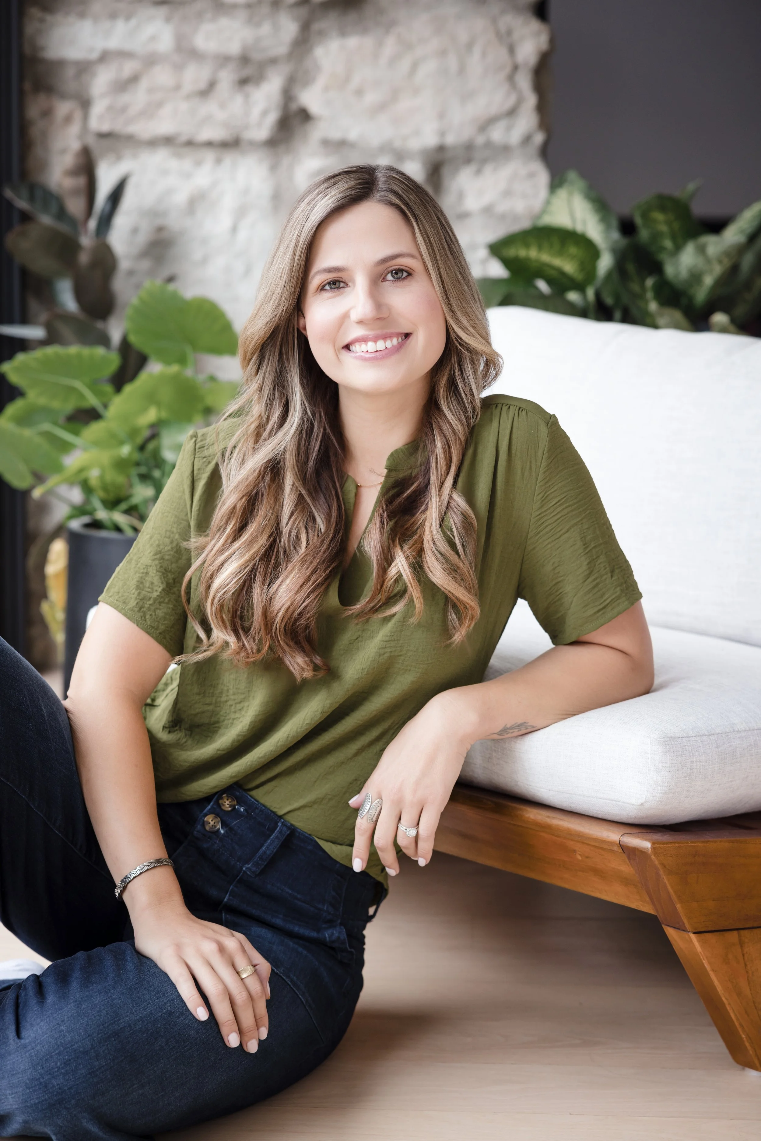 A smiling woman with long wavy hair, wearing a green top and dark jeans, sitting on the floor and leaning against a white sofa in a room with plants and a stone wall background.