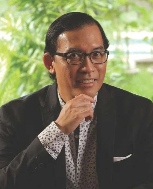 A man with glasses, black hair, wearing a dark blazer and patterned shirt, posing with his hand near his chin outdoors with green foliage in the background.