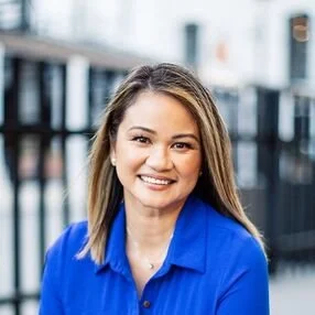 A young woman with long brown hair smiling outdoors, wearing a blue shirt.