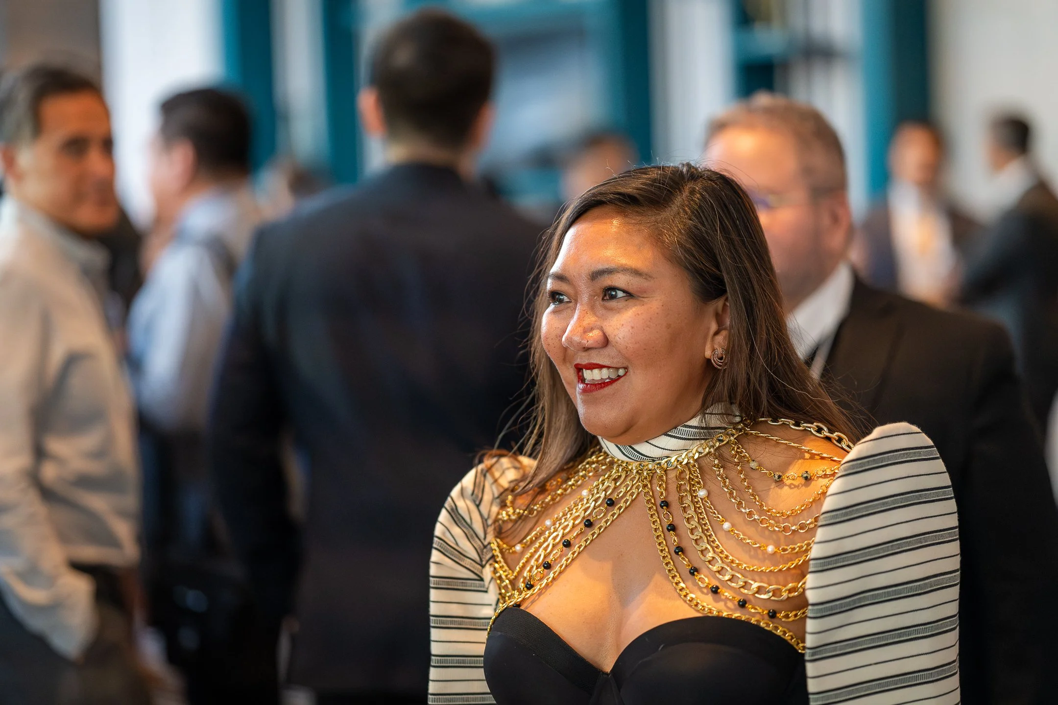 A woman wearing a black dress with a striped blazer and an elaborate gold chain necklace, smiling at a social gathering with people in formal attire in the background.