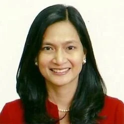 A woman with long black hair smiling, wearing a red top and pearl earrings.
